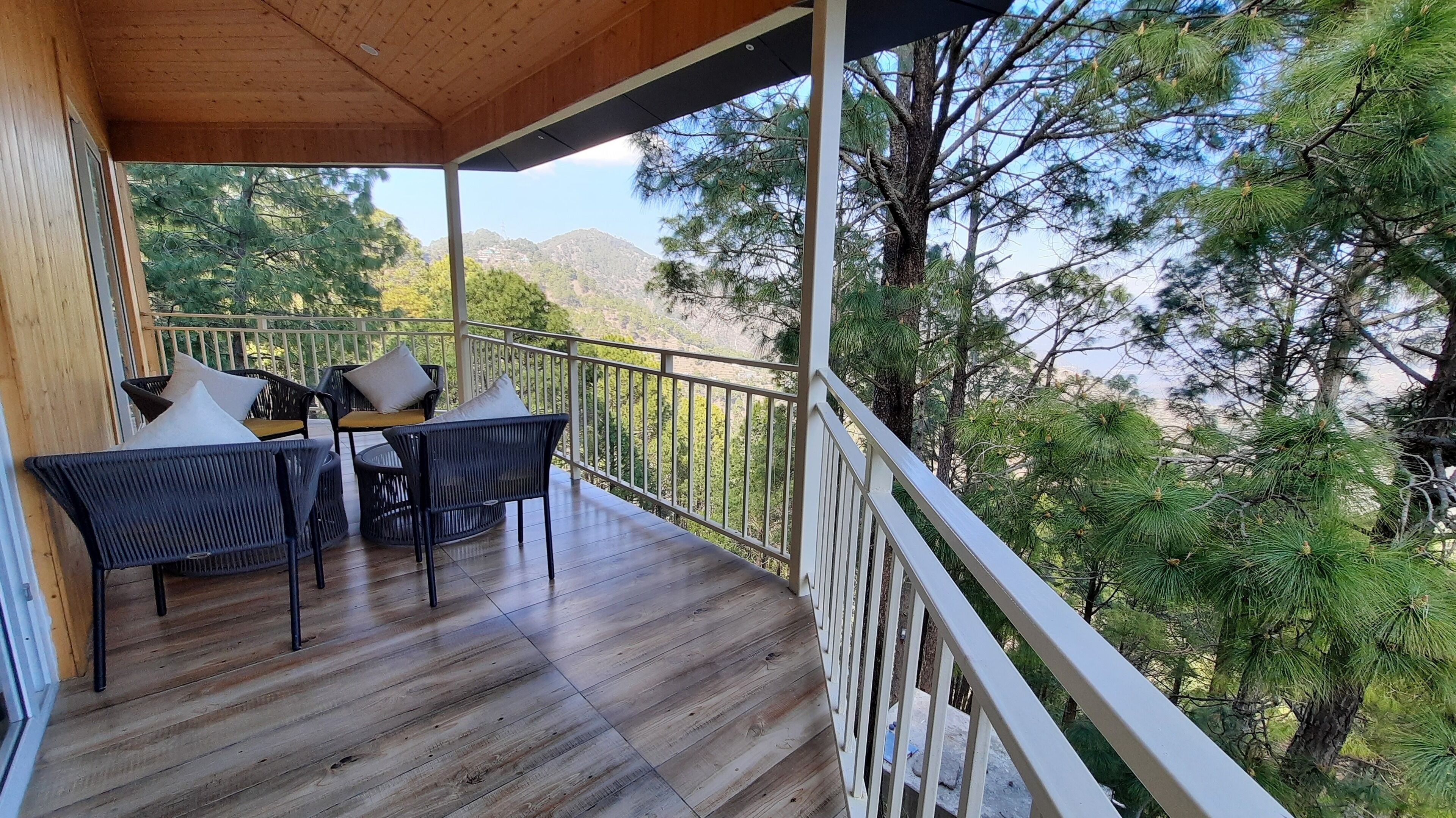 Une terrasse en bois, dotée de mobilier d’extérieur et d’une balustrade blanche, qui offre une vue sur une zone boisée.