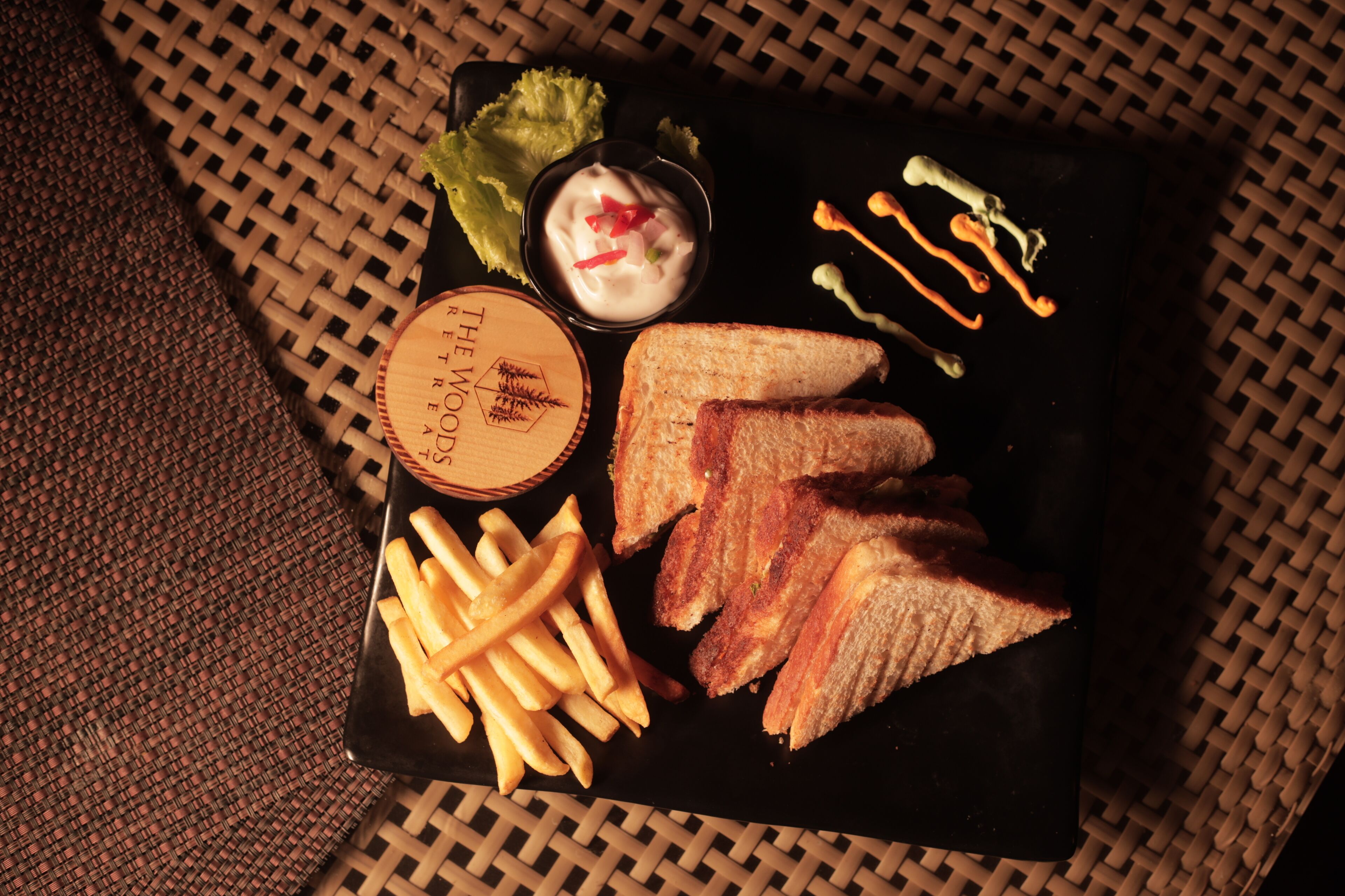 Un plato negro con papas fritas, pan tostado, un recipiente pequeño para salsa y un posavasos de madera con un logotipo.