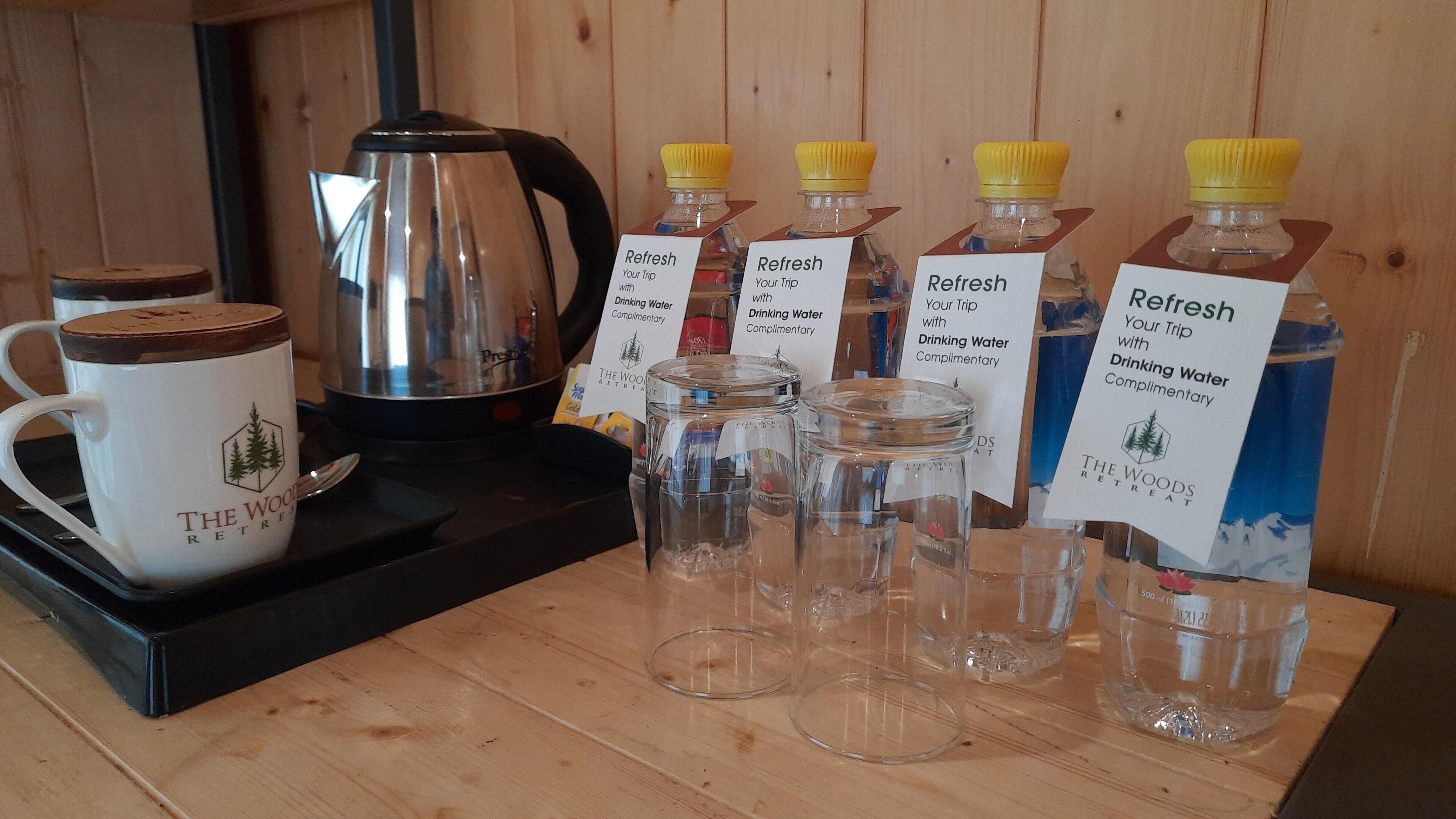 A coffee station with a kettle, mugs, and water bottles labeled "Refresh Your Trip with Drinking Water Complimentary" at The Wood Retreat.