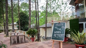 Un panneau indiquant « BIENVENUE AU WOODS RETREAT », placé sur un socle dans une zone boisée, agrémentée de plantes en pot et de sièges d’extérieur.