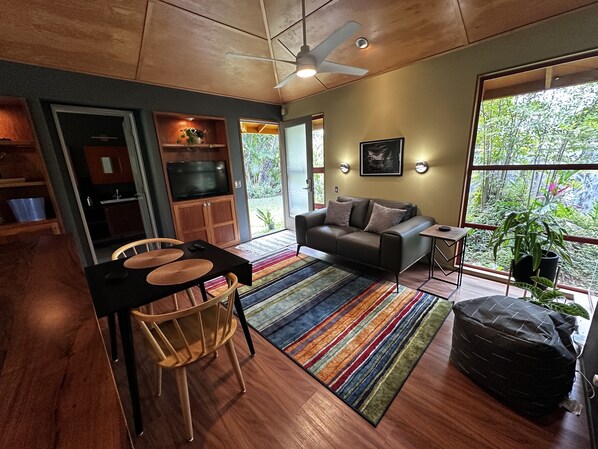 A modern living room with a sofa, a dining table with chairs, a TV, and a large window with a view of greenery. - Tree Top Suite- Beautiful walk to Hanalei Bay (Princeville)
