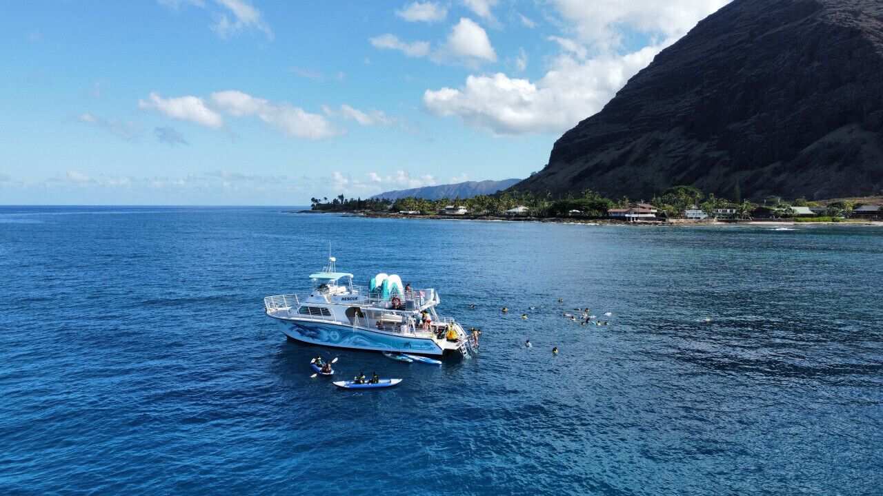 Un grand bateau arborant un panneau indiquant « Sauvetage » à l’avant, entouré d’eaux bleues et calmes et bordé d’une côte montagneuse.