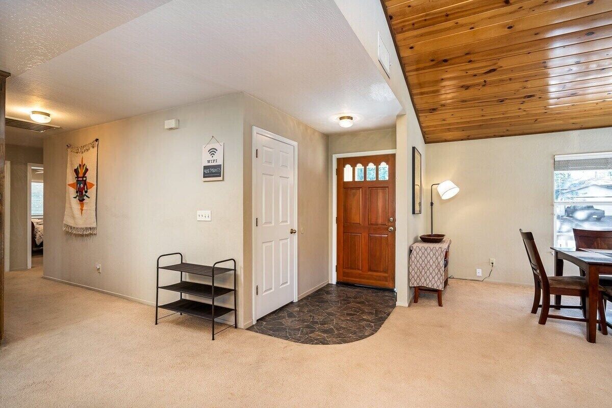 A hallway with a wooden ceiling, a door, a small table with a lamp, and a dining area with chairs.