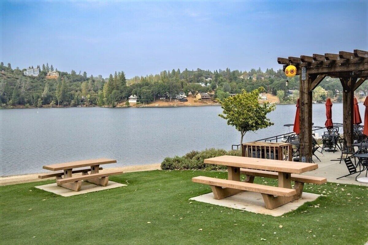 A lakeside picnic area with two picnic tables, a pergola with umbrellas, and a view of a lake and forested hills.