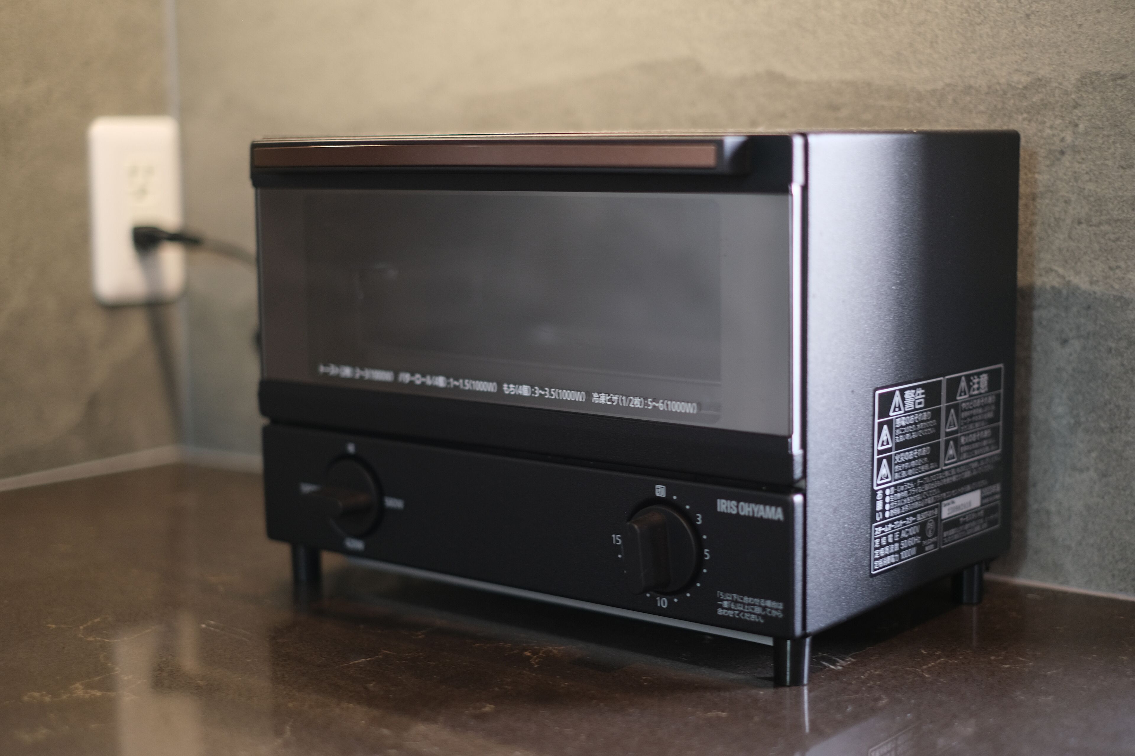 A black electronic device with two knobs and a display screen, placed on a reflective surface against a textured wall.