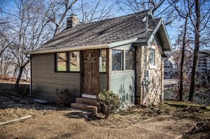 A small, rustic cabin with a wooden door and windows, surrounded by trees and a stone wall.