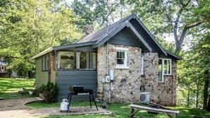 Een klein stenen huis met een schuin dak, een voortuin en een buitenbarbecue.