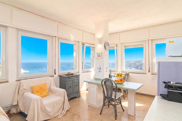 A room with a view of the sea, a dining area with a table and chairs, a sofa, and a small kitchen area.