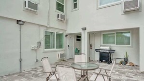 A patio with a round glass table and four white chairs, a grill, and a potted plant. - Bright Studio | Walk to Pompano Beach & Pier (Pompano Beach)