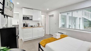 A hotel room with a bed, a desk, a kitchenette, and a large window with blinds. - Bright Studio | Walk to Pompano Beach & Pier (Pompano Beach)