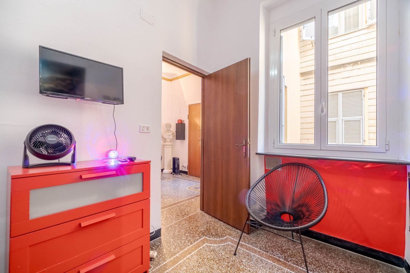 A room with a TV mounted on the wall, a red dresser, a chair, and a window with blinds.
