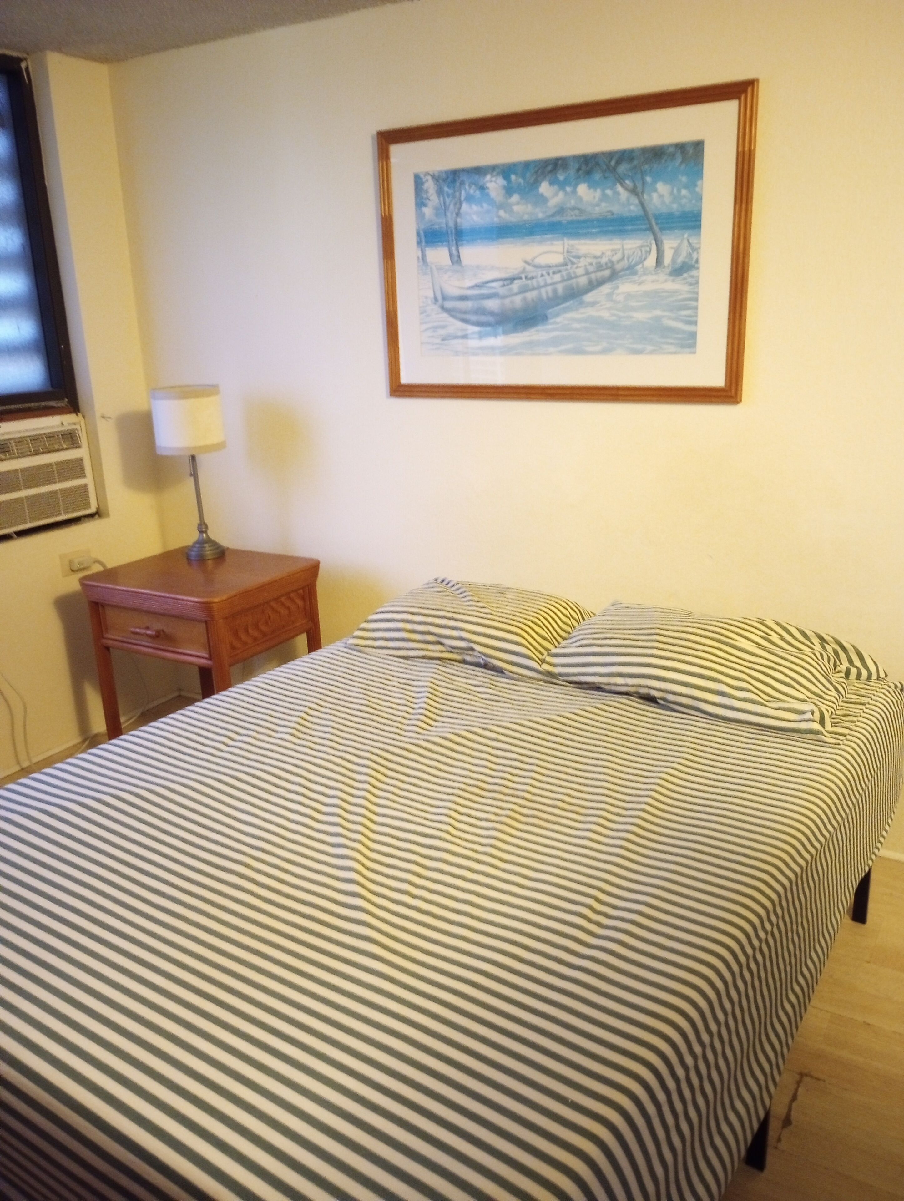 Una cama con ropa de cama a rayas, una mesita de noche con una lámpara, un cuadro en la pared y una ventana con aire acondicionado.