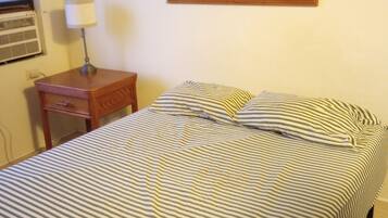 Una cama con ropa de cama a rayas, una mesita de noche con una lámpara, un cuadro en la pared y una ventana con aire acondicionado.