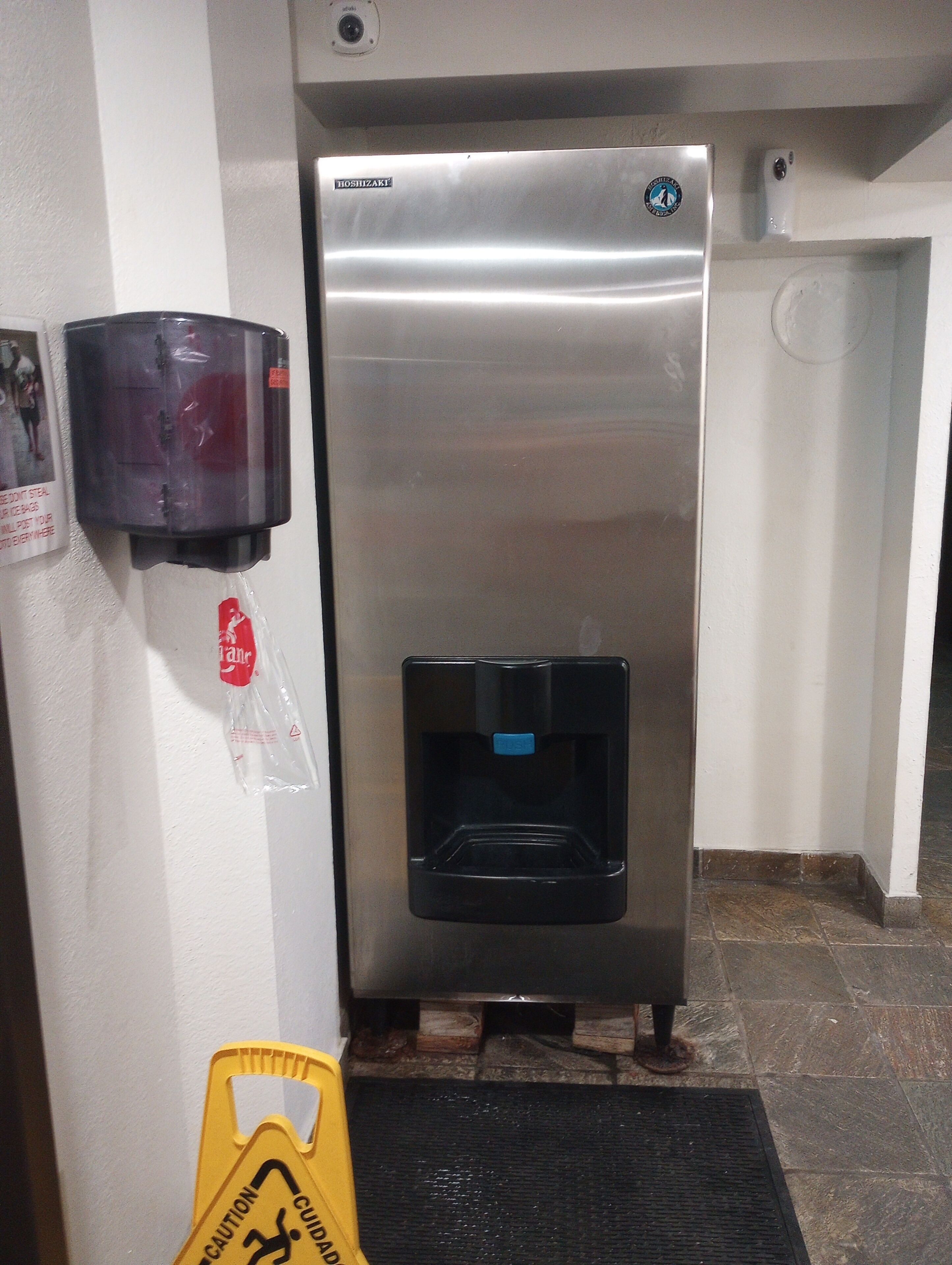 Un refrigerador de acero inoxidable con dispensador de agua, una cámara de seguridad y un letrero de precaución en el suelo.