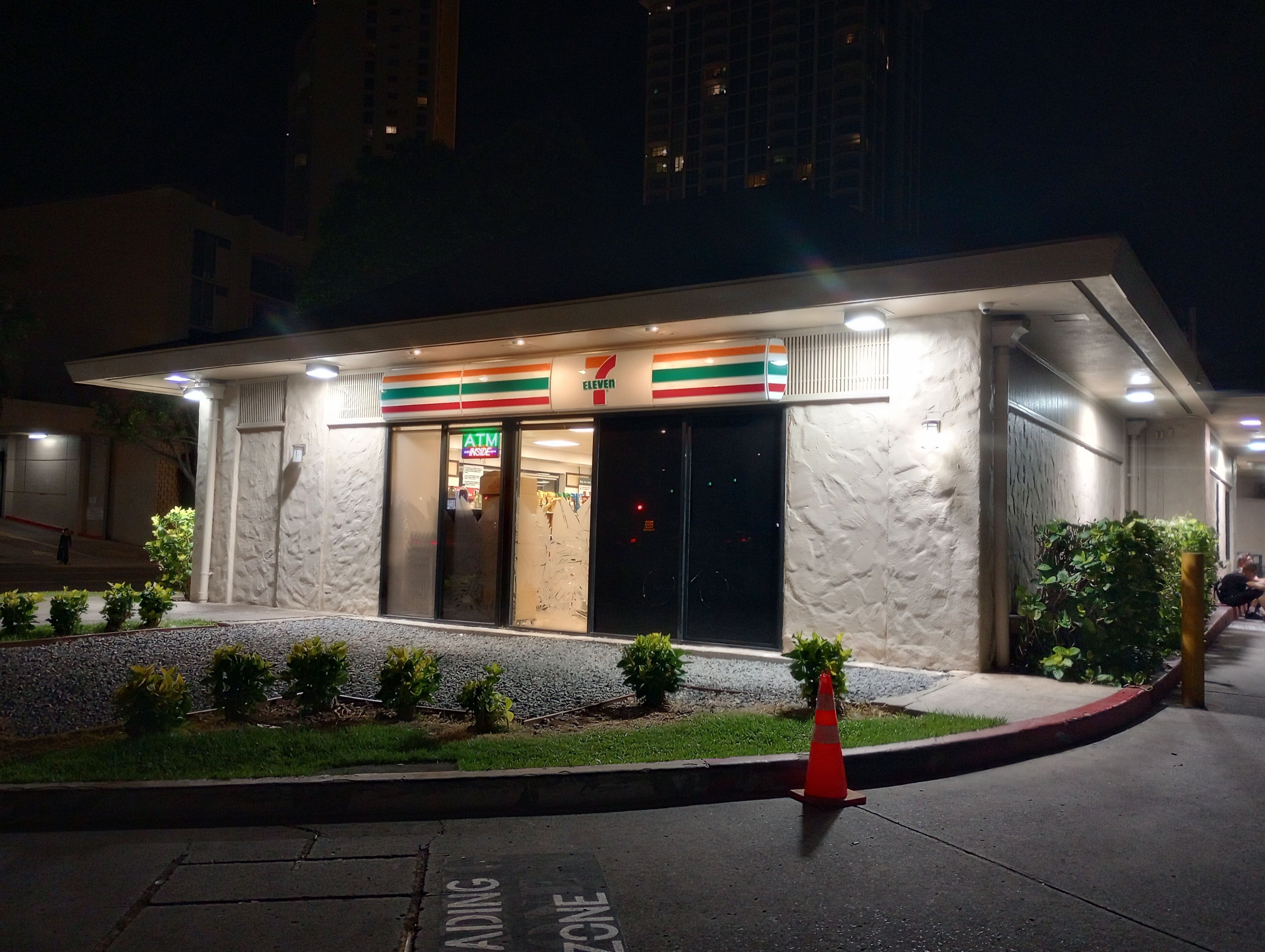 Una tienda 7-Eleven de noche, con fachada de piedra, un pequeño jardín y un estacionamiento.