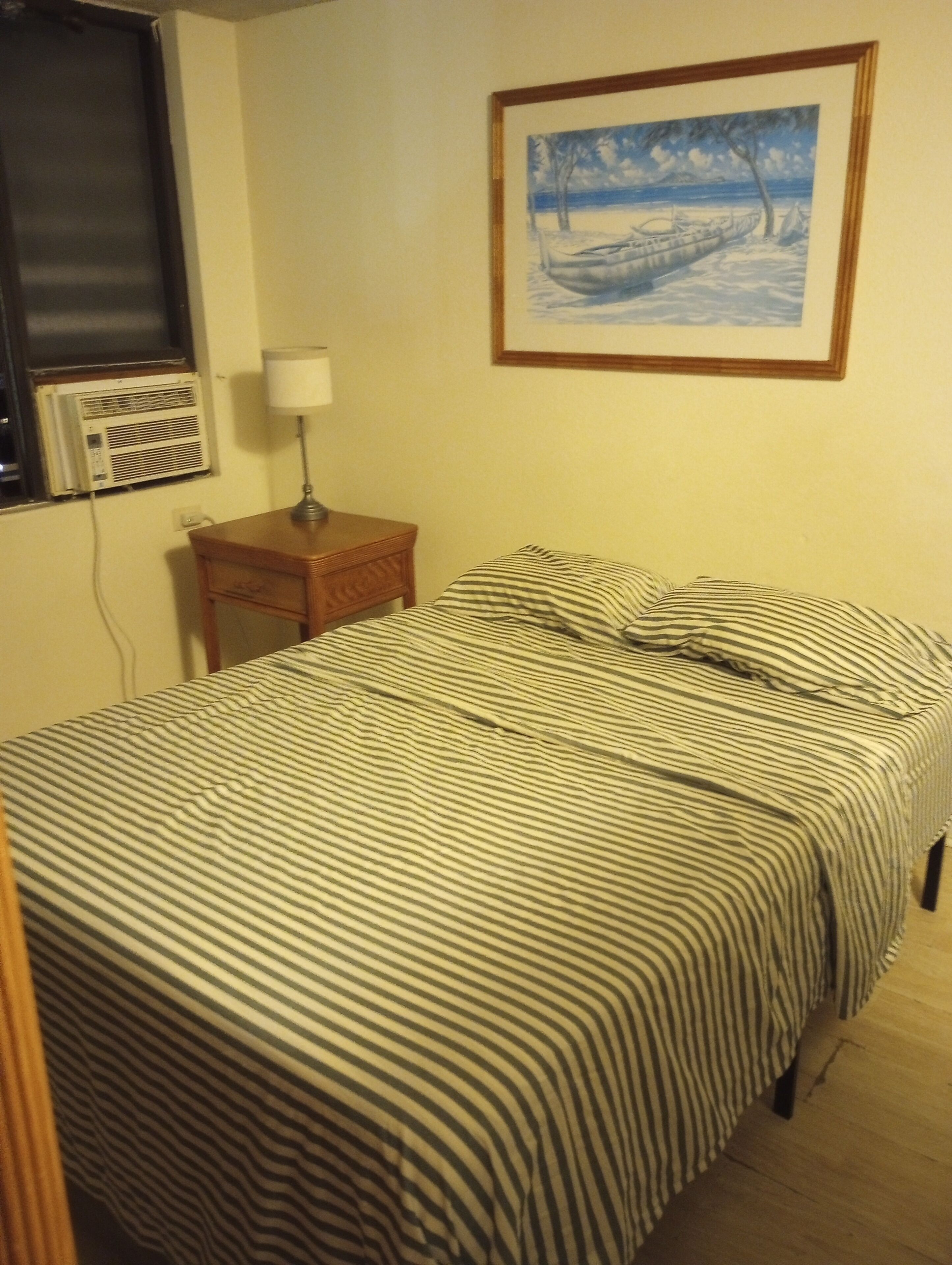 Una cama con ropa de cama a rayas, una mesita de noche con una lámpara, un cuadro enmarcado en la pared y un equipo de aire acondicionado.