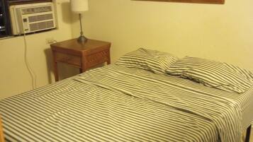 Una cama con ropa de cama a rayas, una mesita de noche con una lámpara, un cuadro enmarcado en la pared y un equipo de aire acondicionado.