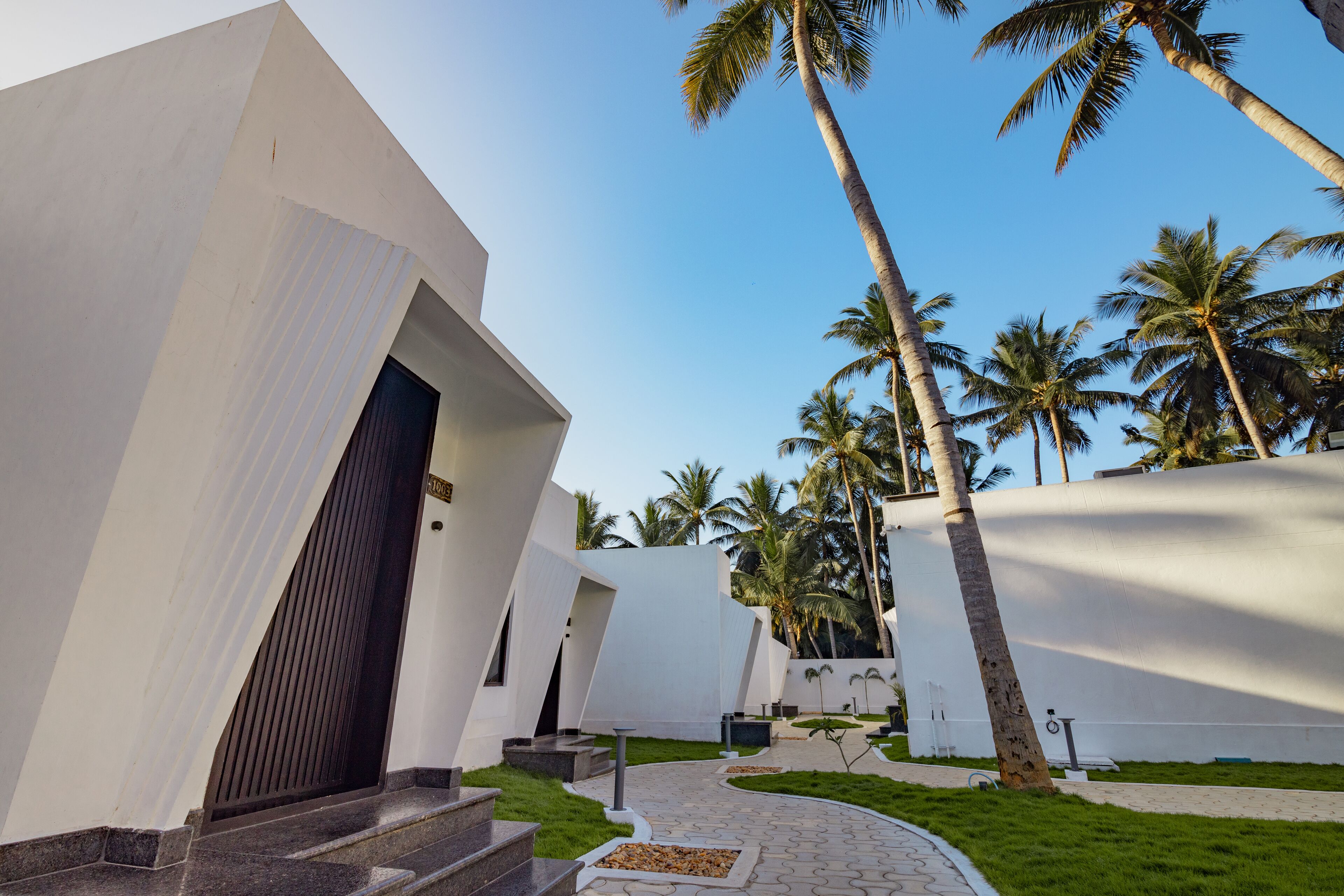 A modern building with a curved facade, a pathway leading to a door, and palm trees in the background.