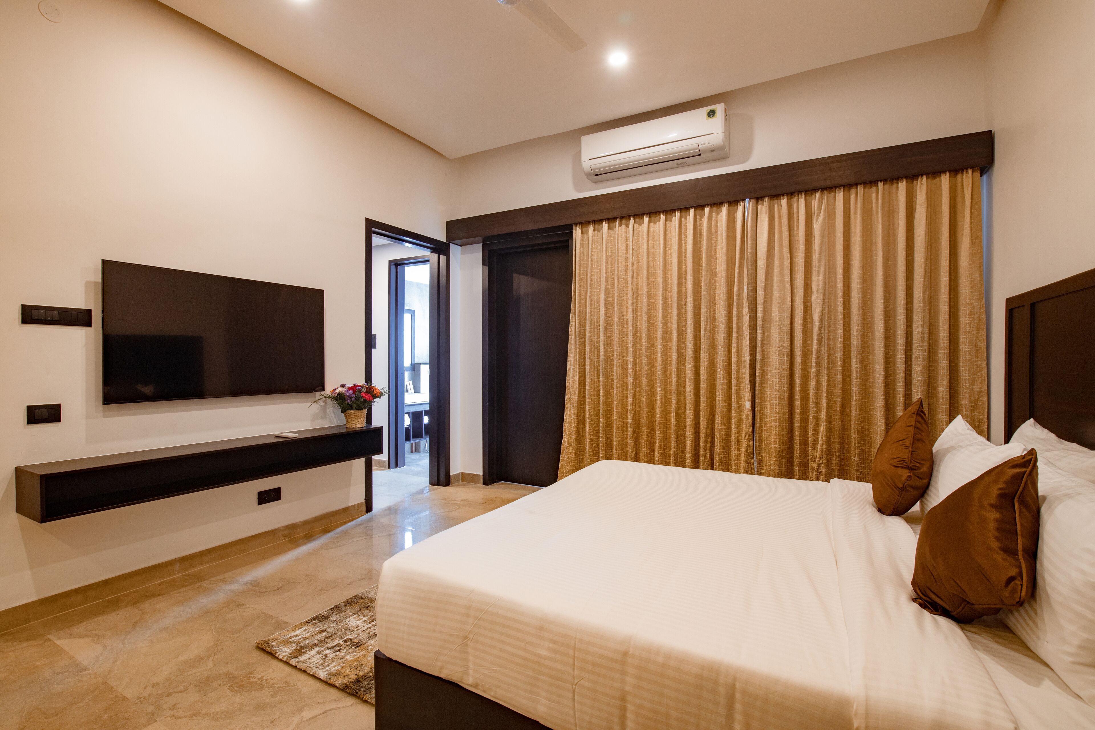 A modern hotel room with a large bed, a flat-screen TV mounted on the wall, a built-in air conditioning unit, and a sliding door leading to a bathroom.