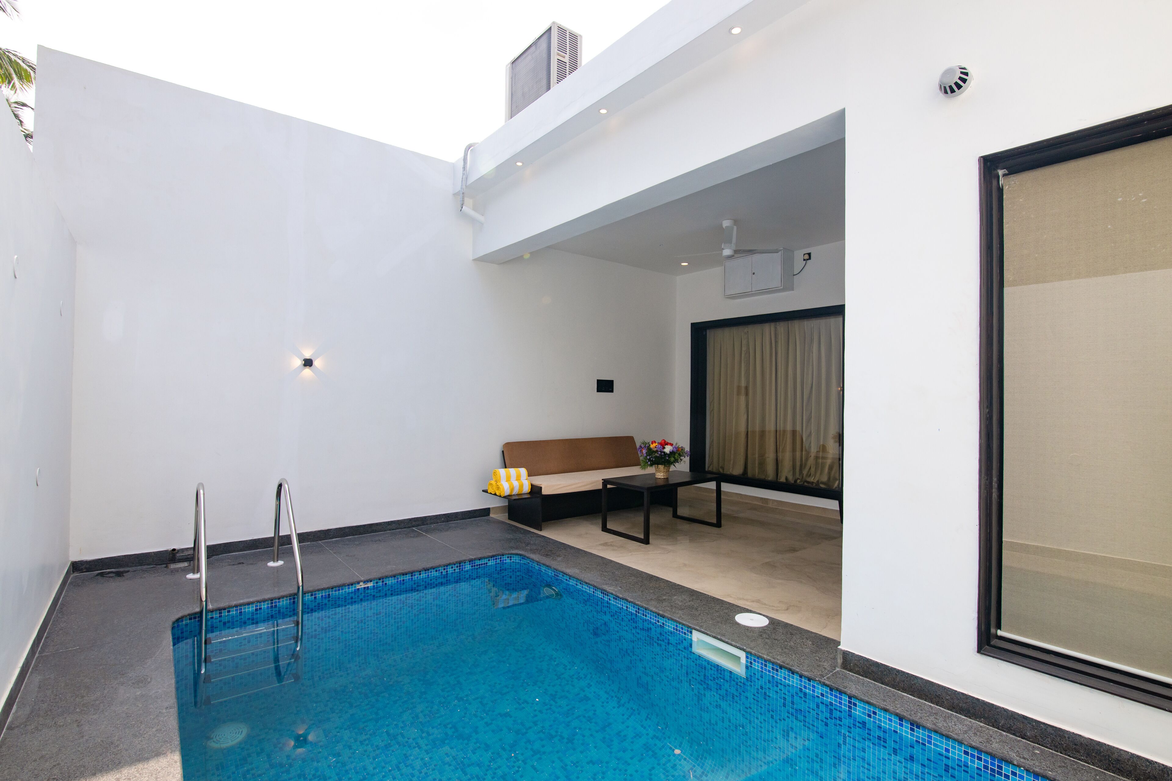 A modern indoor swimming pool with a clear blue water, a small seating area with a sofa and table, and a large window with curtains.