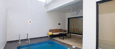 A modern indoor swimming pool with a clear blue water, a small seating area with a sofa and table, and a large window with curtains.