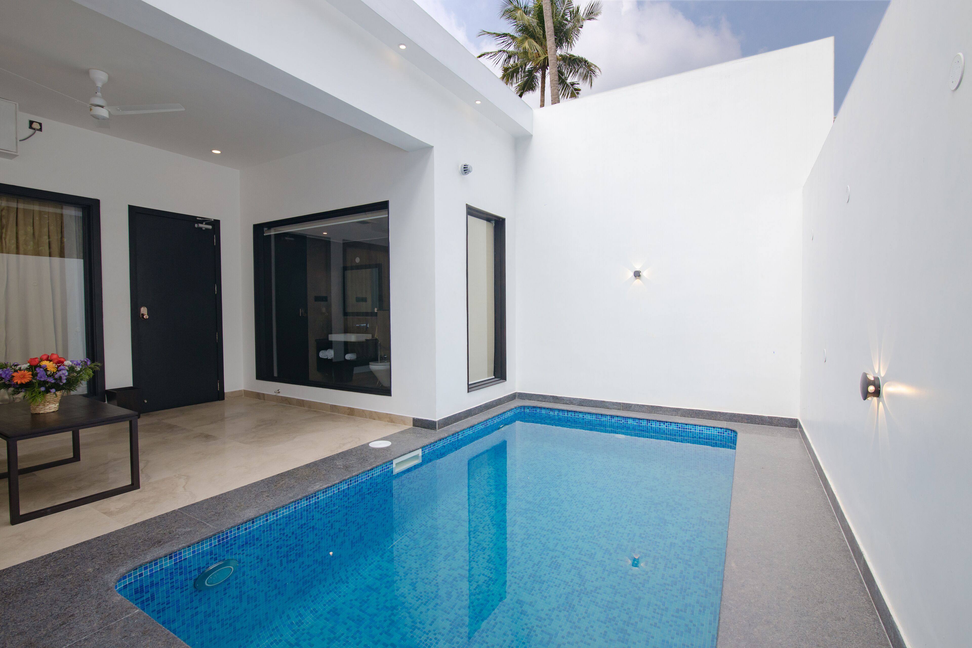 A modern indoor swimming pool with clear blue water, surrounded by white walls and a ceiling fan.