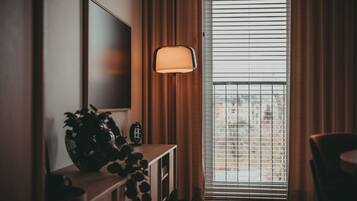 A room with a TV mounted on the wall, a floor lamp, a plant on a console table, and a window with blinds.
