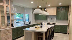 Une cuisine moderne avec des meubles de couleur verte, un grand îlot central, des luminaires suspendus et une vue sur l’extérieur.