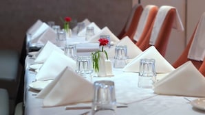 A long dining table set with white napkins, clear glasses, and a small vase with a red flower.