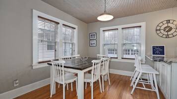 A dining area with a wooden table and white chairs, large windows with blinds, a wall clock, and a welcome sign.