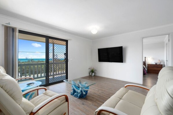 A spacious living room with a balcony, a glass coffee table, and a flat-screen TV mounted on the wall.