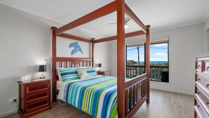 A four-poster bed with a colorful striped bedspread, a nightstand with a lamp, a wooden dresser, and a balcony with a view. - Escape to Paradise - Beach views 5bedrooms, 2 Kitchens, 2 Living, 2 Bathrooms (Paradise Beach)