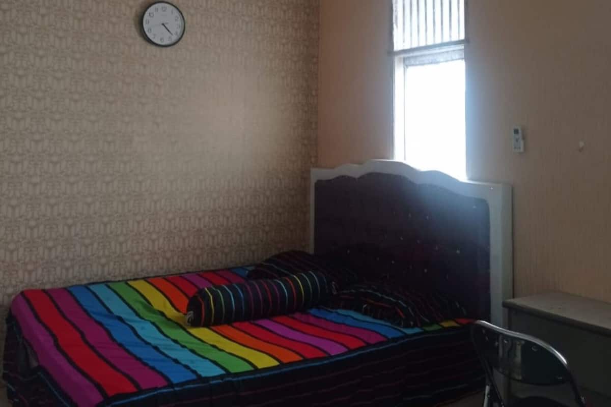 Een kleine eenpersoonskamer met een gestreept dekbed, een klok aan de muur en een raam met jaloezieën.