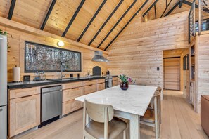 Une cuisine moderne aménagée dans une cabane en rondins, avec un îlot central, des appareils en acier inoxydable et une grande fenêtre donnant sur une forêt.