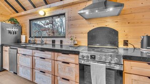 A modern kitchen with wooden walls, a stainless steel stove, and a large window overlooking a forest.