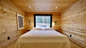 A wooden cabin bedroom with a large bed, a window with a view of trees, and a framed leaf artwork on the wall.