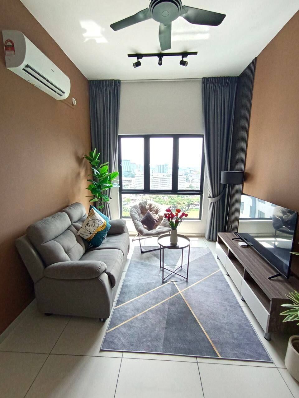 Un salon moderne avec un canapé gris, une petite table basse, un téléviseur posé sur un meuble en bois, un ventilateur de plafond et une grande fenêtre avec des rideaux.