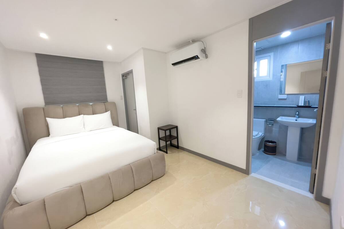A modern bedroom with a large bed, a small bedside table, and a bathroom visible through a glass door.