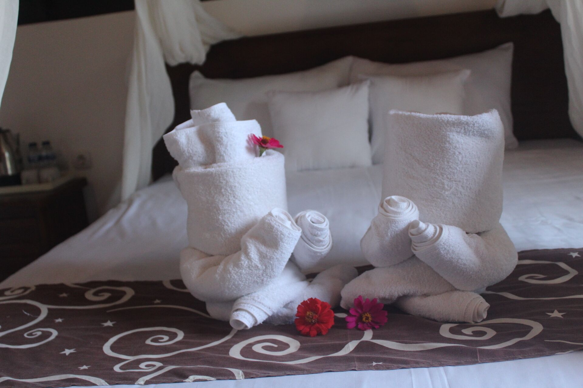 Ein Bett mit weißen Handtüchern, die wie menschliche Figuren geformt sind, einer braunen, gemusterten Bettdecke und zwei roten Blumen auf dem Bett.