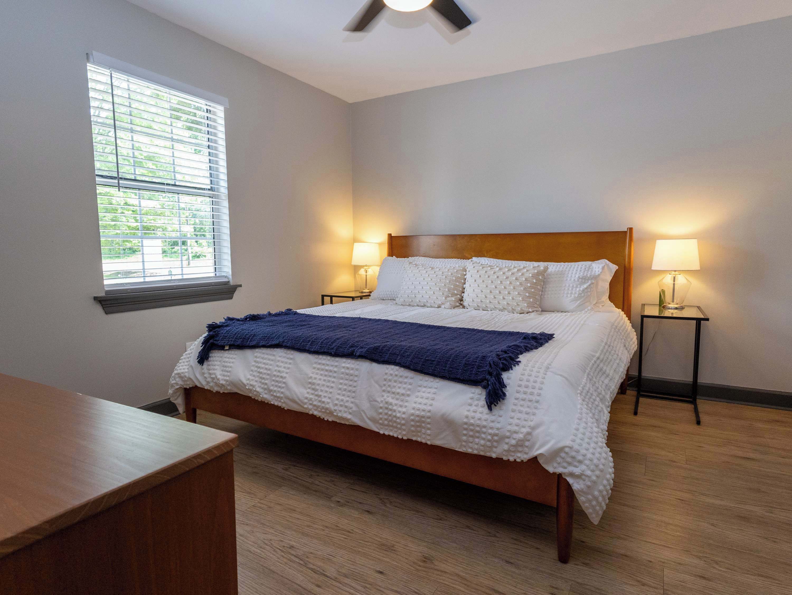 A bedroom with a bed, two bedside lamps, a window with blinds, and a ceiling fan.