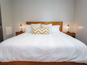 A neatly made bed with a textured white duvet, a wooden headboard, and two bedside lamps.