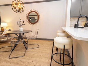 A modern kitchen with a round dining table, clear chairs, a bar stool, a pendant light, and a mirror on the wall.