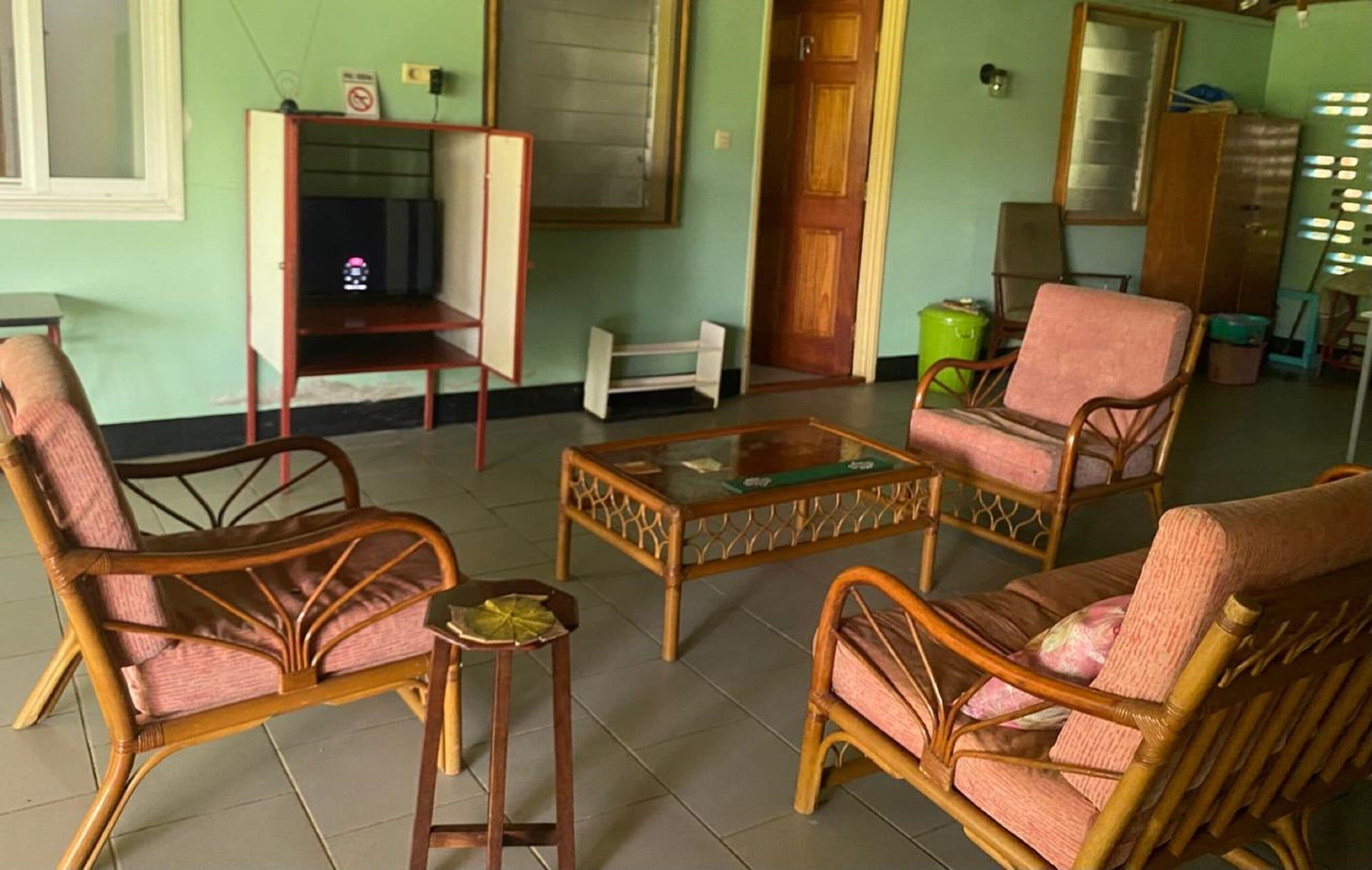 Quarto com mobiliário de vime, uma mesa de centro de vidro e uma televisão sobre um suporte.