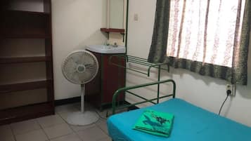A room with a bed, a bedside table, a fan, a sink, and a window with curtains.