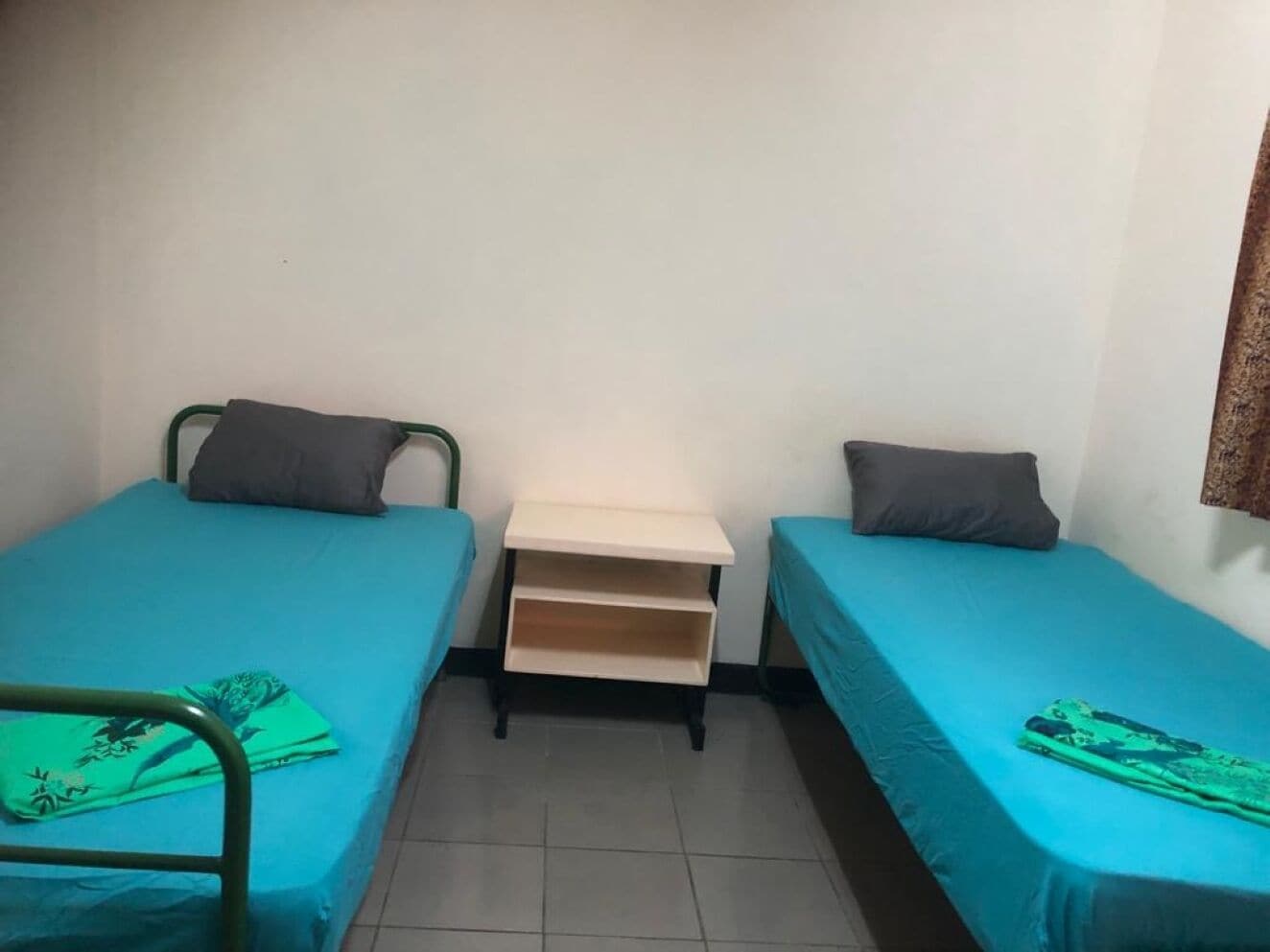 A small room with two single beds, each with a blue bedspread and a pillow. There is a small wooden nightstand with a shelf between the beds.