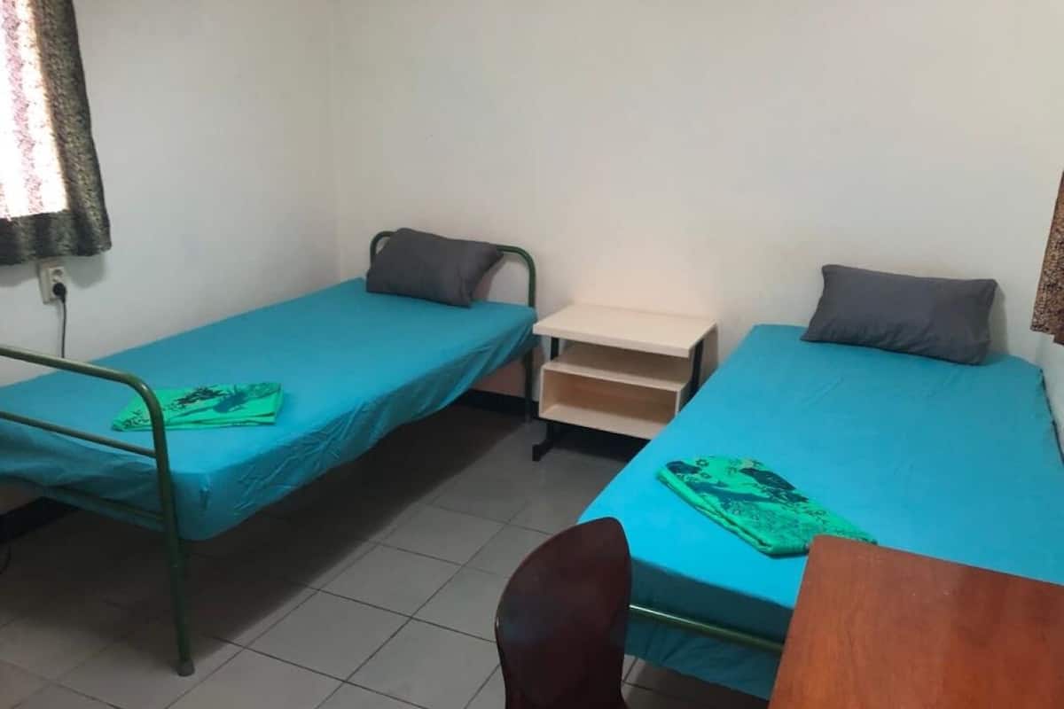 A dormitory room with two single beds, each with a pillow and a green blanket. There is a wooden desk and a chair in the foreground.