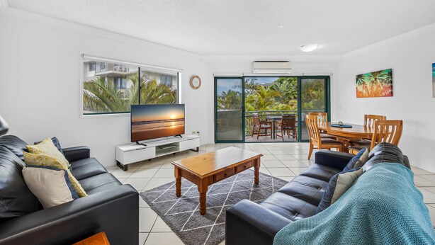 A modern living room with a TV, leather sofas, a wooden coffee table, and a balcony with a dining area.