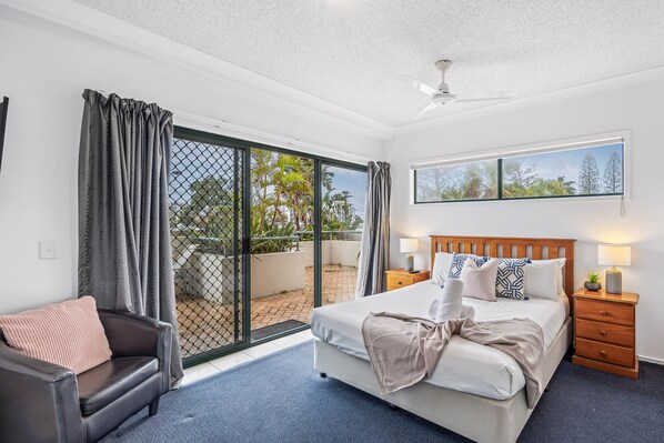 A hotel room with a bed, a nightstand, a chair, a window with a view of trees, and a ceiling fan.