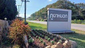 Un letrero con escritura tailandesa, un jardín ajardinado con rocas y plantas, y una carretera al fondo.