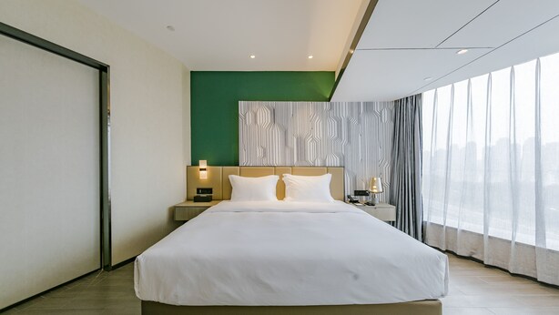 Une chambre d’hôtel moderne dotée d’un grand lit, d’une table de chevet avec une lampe, d’un mur d’accent vert et offrant une vue sur la ville grâce à de grandes fenêtres.